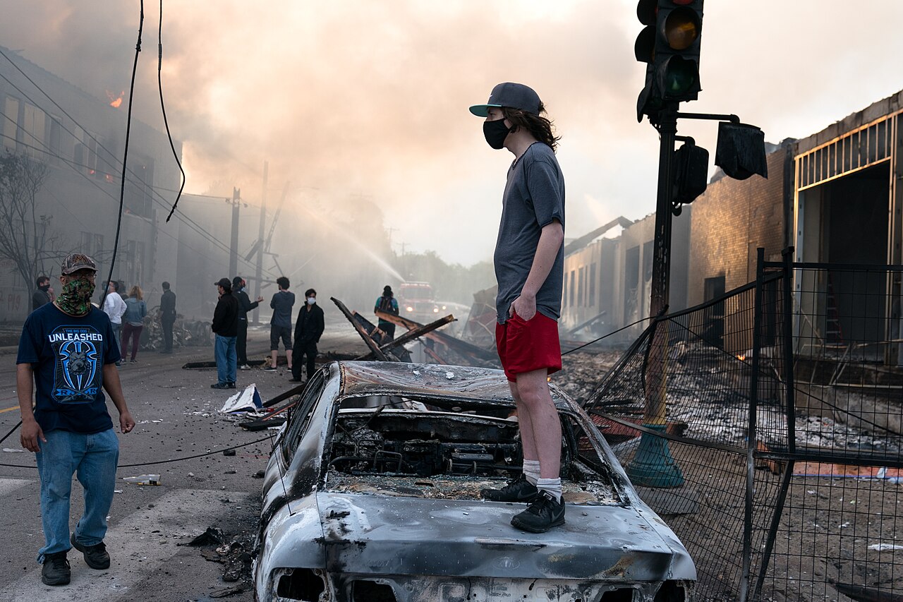 A man stands on a burned out car on Thursday morning as fires burn behind him in the Lake St area of Minneapolis, Minnesota (49945886467)